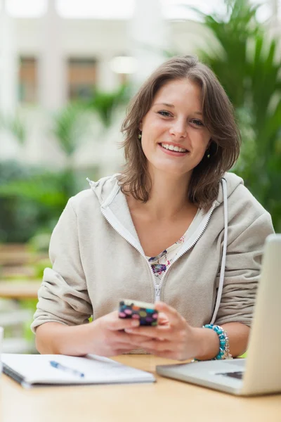 Kvinnlig student med mobiltelefon och bärbar dator vid cafeterian bord — Stockfoto