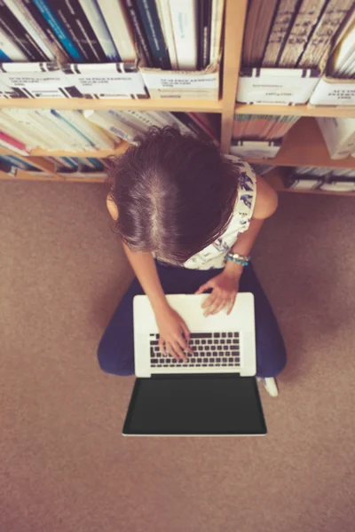 Student tegen boekenplank met behulp van laptop op de verdieping van de bibliotheek — Stockfoto