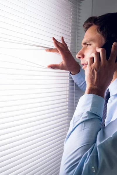 Businessman peeking through blinds while on call in office — Stock Photo, Image
