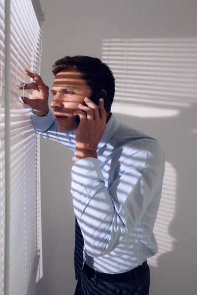 Businessman peeking through blinds while on call in office — Stock Photo, Image