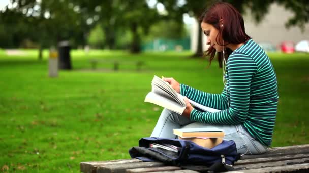 Estudiante sentado en el banco leyendo un libro escuchando música — Vídeo de stock