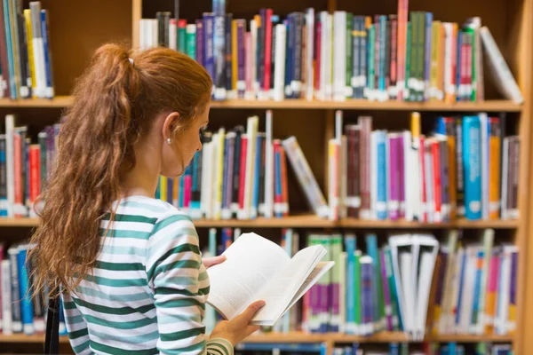 Рыжая студентка читает книгу с полки в библиотеке — стоковое фото