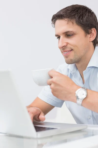 Leende ung man med tekopp med laptop hemma — Stockfoto