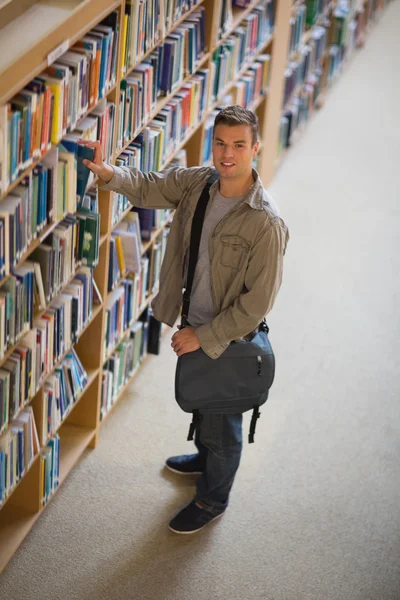 Студент бере книгу з полиці в бібліотеці, посміхаючись на камеру — стокове фото