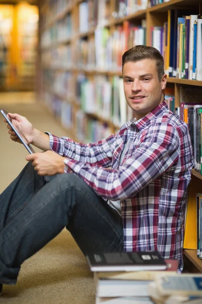 Souriant jeune étudiant assis sur le sol de la bibliothèque en utilisant la tablette — Photo