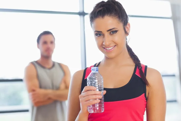 Fit mujer sosteniendo la botella de agua con un amigo en el fondo en el gimnasio —  Fotos de Stock