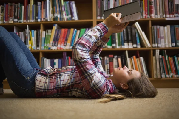 Красивая студентка лежит на полу библиотеки и читает книгу. — стоковое фото