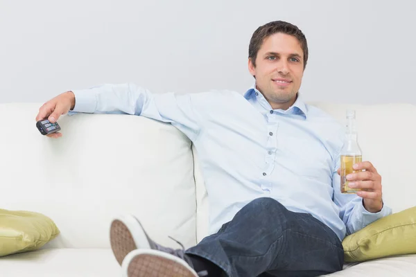 Hombre con una bebida y control remoto sentado en el sofá en casa — Foto de Stock
