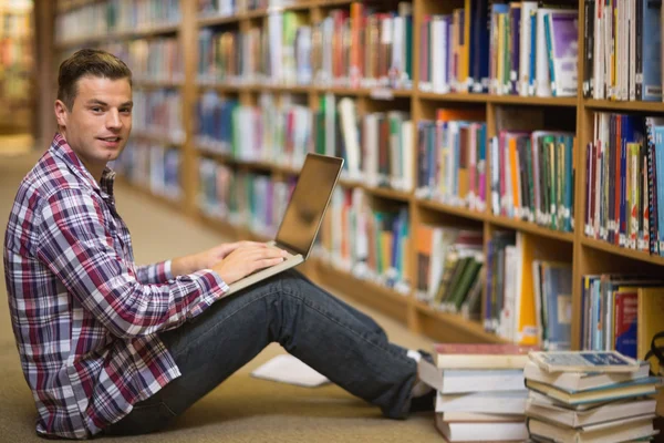 Estudiante joven serio sentado en el suelo de la biblioteca usando el ordenador portátil —  Fotos de Stock