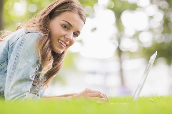 Glückliche Studentin, die mit ihrem Laptop im Gras liegt und in die Kamera schaut — Stockfoto