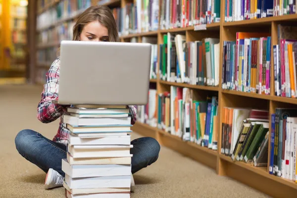 Young student sitting on library floor using laptop on pile of books — Stock Photo, Image