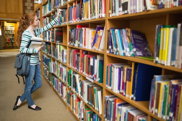 Рыжая студентка берет книгу с полки в библиотеке — стоковое фото