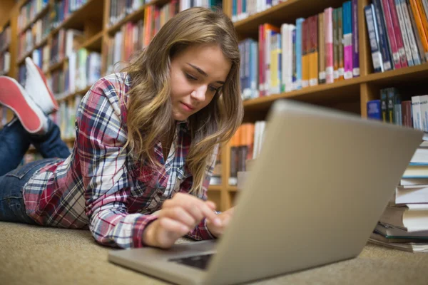 Jovem estudante feliz deitado no chão da biblioteca usando laptop — Fotografia de Stock