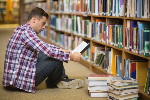 Красивый молодой студент, сидящий на полу библиотеки и читающий книгу — стоковое фото