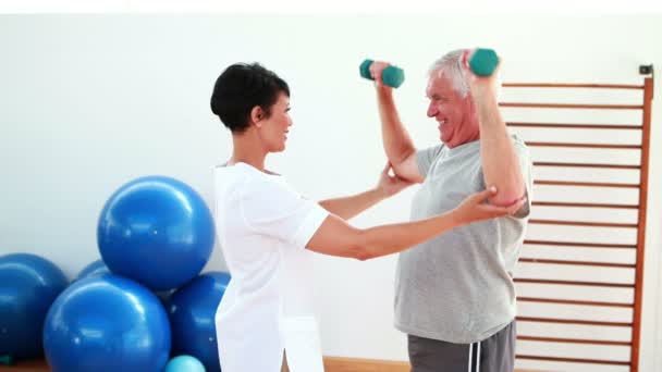 Pretty physiotherapist helping elderly patient lift hand weights — Stock Video