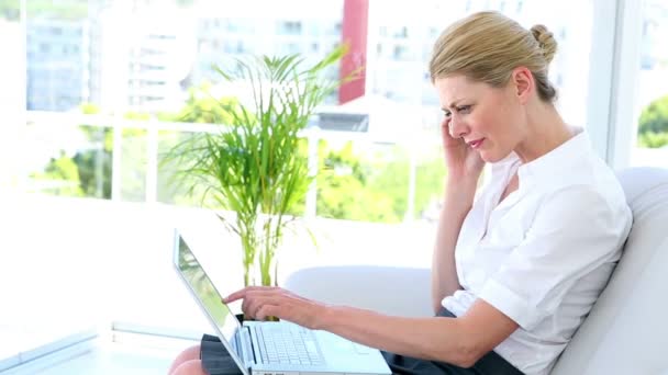 Бізнес-леді сидить на дивані, використовуючи ноутбук розмовляючи по телефону — стокове відео
