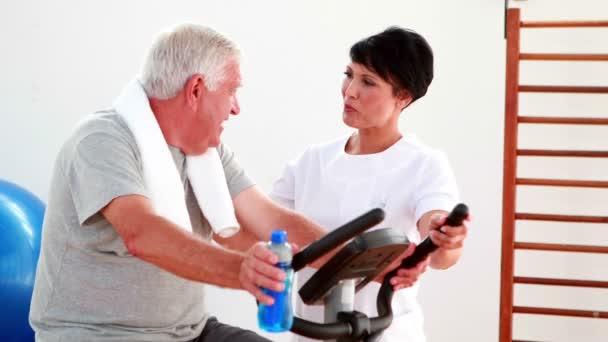 Hombre mayor usando la bicicleta estática hablando con el fisioterapeuta — Vídeos de Stock