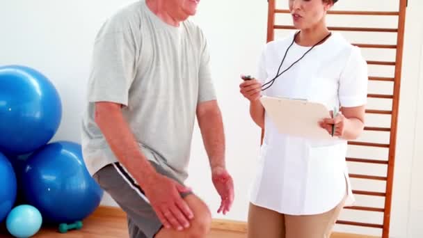Smiling fisioterapeuta cronometrar paciente idoso com joelho dobrado — Vídeo de Stock