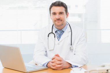 Male doctor with laptop at desk in medical office clipart