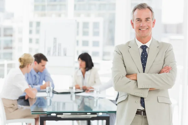 Happy businessman looking at camera while staff discuss behind him — Stock Photo, Image