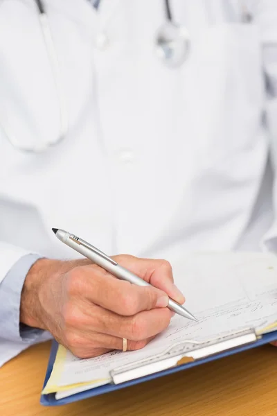 Doctor sentado en su escritorio escribiendo en el portapapeles —  Fotos de Stock