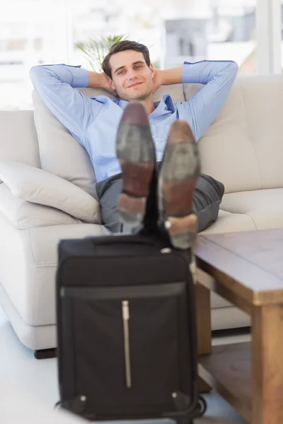 Smiling businessman sitting on couch with feet up on suitcase — Stock Photo, Image