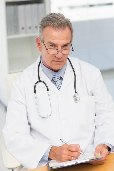 Serious doctor sitting at his desk writing on clipboard — Stock Photo, Image