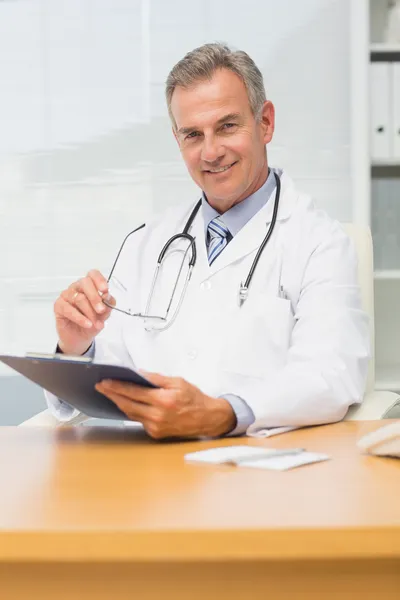 Leende läkare sitter vid sitt skrivbord med Urklipp — Stockfoto