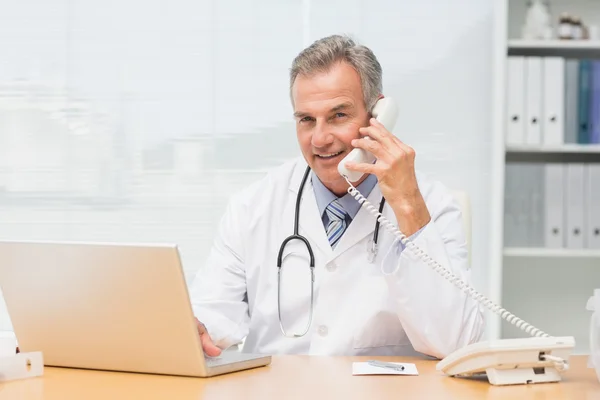 Médico sorrindo usando laptop e falando no telefone na mesa — Fotografia de Stock