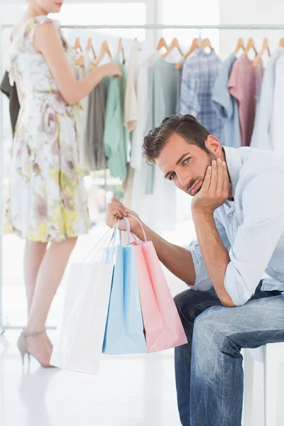 Homem entediado com sacos de compras, enquanto a mulher pelo rack de roupas — Fotografia de Stock