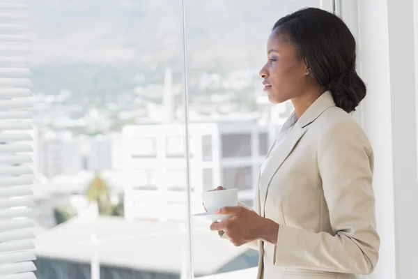 Femme d'affaires élégante avec tasse à thé regardant par la fenêtre du bureau — Photo