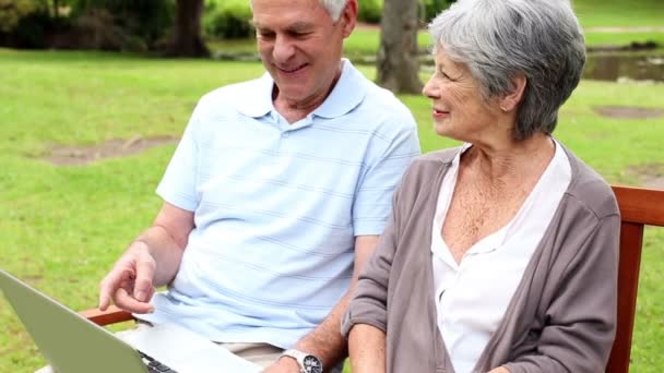 Пенсионная пара, сидящая на скамейке в парке с ноутбуком — стоковое видео