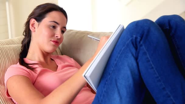 Pretty brunette writing in notepad on the sofa — Stock Video