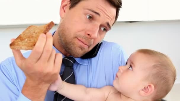 Empresario sosteniendo a su bebé y hablando por teléfono mientras come tostadas — Vídeos de Stock