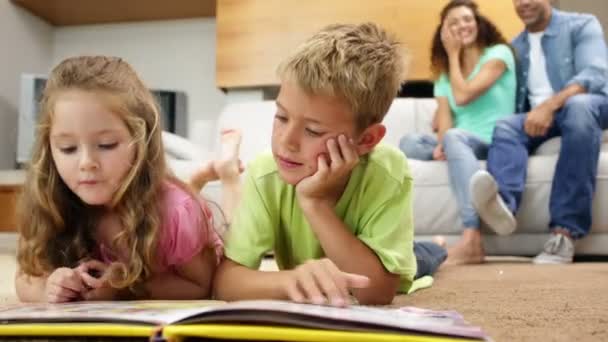 Hermanos tumbados en el suelo leer libro con los padres detrás de ellos en el sofá — Vídeo de stock