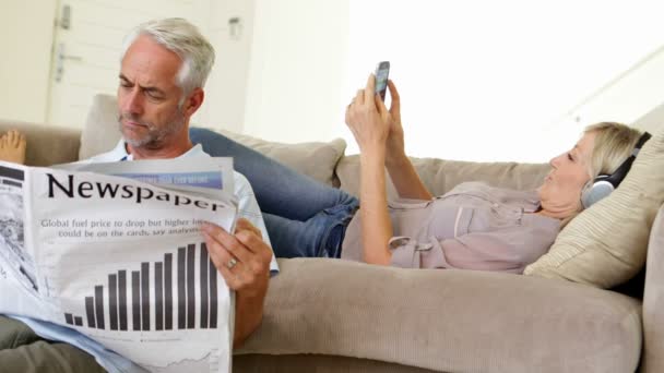 Человек читает бумагу, пока партнер слушает музыку на диване — стоковое видео