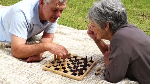 Seniorenpaar entspannt im Park auf einer Decke liegend Schach spielend — Stockvideo