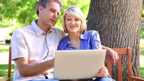 Ласковая пара, сидящая на скамейке в парке с ноутбуком — стоковое видео