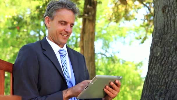 Biznesmen, siedząc na ławce w parku za pomocą tabletki — Wideo stockowe