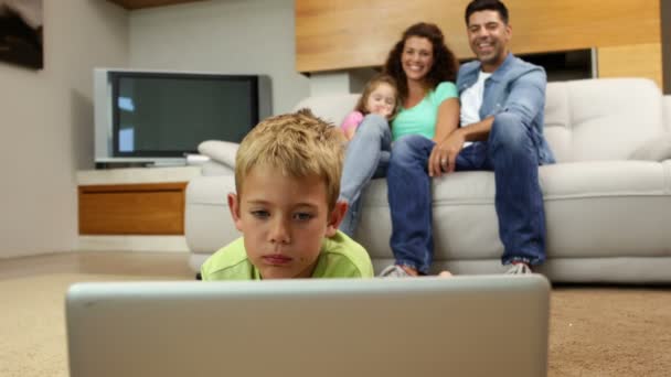 Kleine jongen liggend op vloer laptop met familie achter hem op sofa — Stockvideo