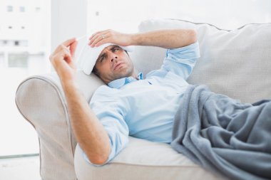 Sick man lying on sofa checking his temperature under a blanket clipart