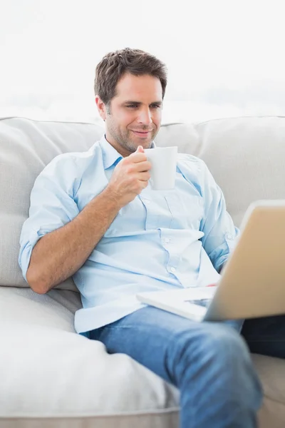 Hombre contento usando el portátil sentado en el sofá tomando un café — Foto de Stock