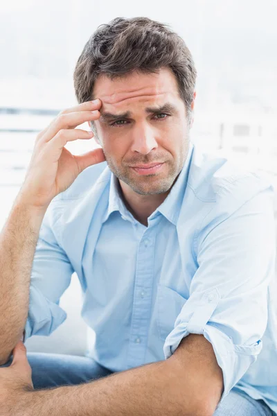 Wincing man with headache sitting on the couch looking at camera — Stock Photo, Image