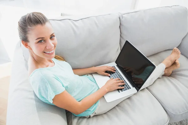 Ευτυχισμένη γυναίκα που κάθεται στον καναπέ, χρησιμοποιώντας την lap-top χαμογελά στη φωτογραφική μηχανή — Φωτογραφία Αρχείου