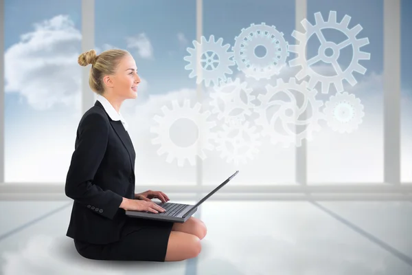 Blonde businesswoman sitting using laptop with cogs and wheels — Stock Photo, Image