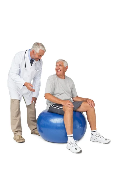 Мужской терапевт, глядя на старшего мужчину, сидит на мяче для упражнений — стоковое фото