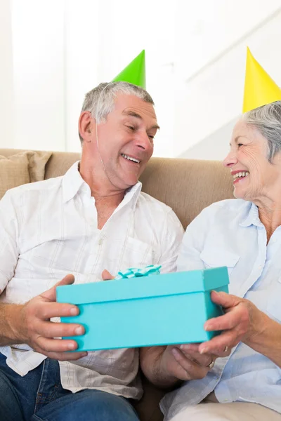 Äldre par sitter på soffan klädd partyhattar innehar en gåva — Stockfoto