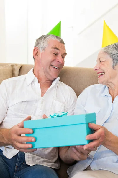 Pareja mayor sentada en un sofá con sombreros de fiesta sosteniendo un regalo — Foto de Stock