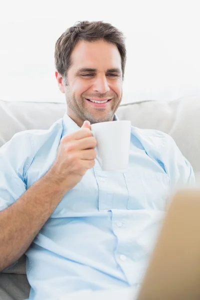 Hombre sonriente usando un portátil sentado en un sofá tomando un café —  Fotos de Stock