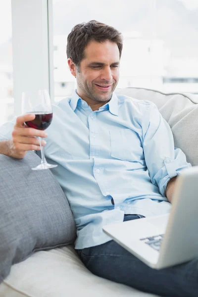 Homem feliz relaxando no sofá com copo de vinho tinto usando laptop — Fotografia de Stock